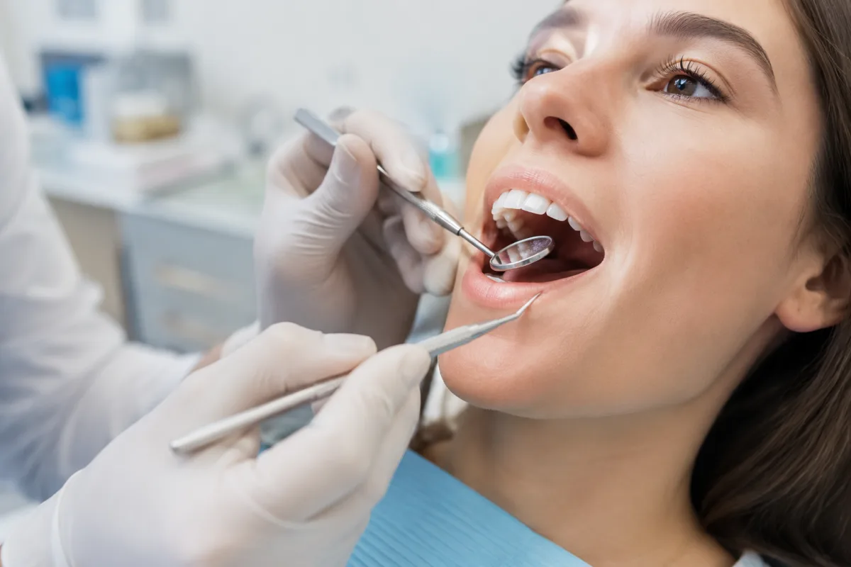 Ce que coûte vraiment le dentiste en Belgique (et ce que rembourse la mutuelle) Main gantée d'un dentiste tenant un miroir buccal devant la bouche ouverte d'un patient allongé